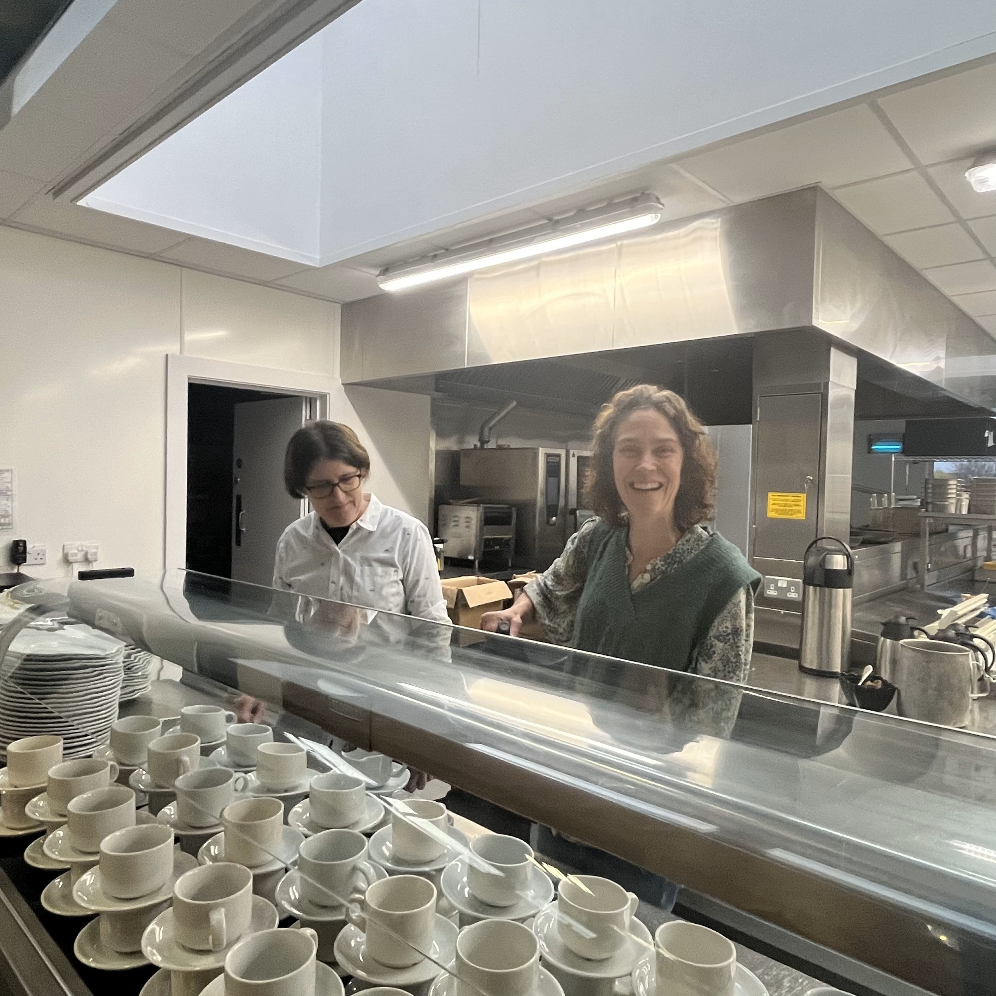 Picture of two women behind a canteen counter serving drinks with lots of cups and saucers in front of them. There is a large school kitchen behind them