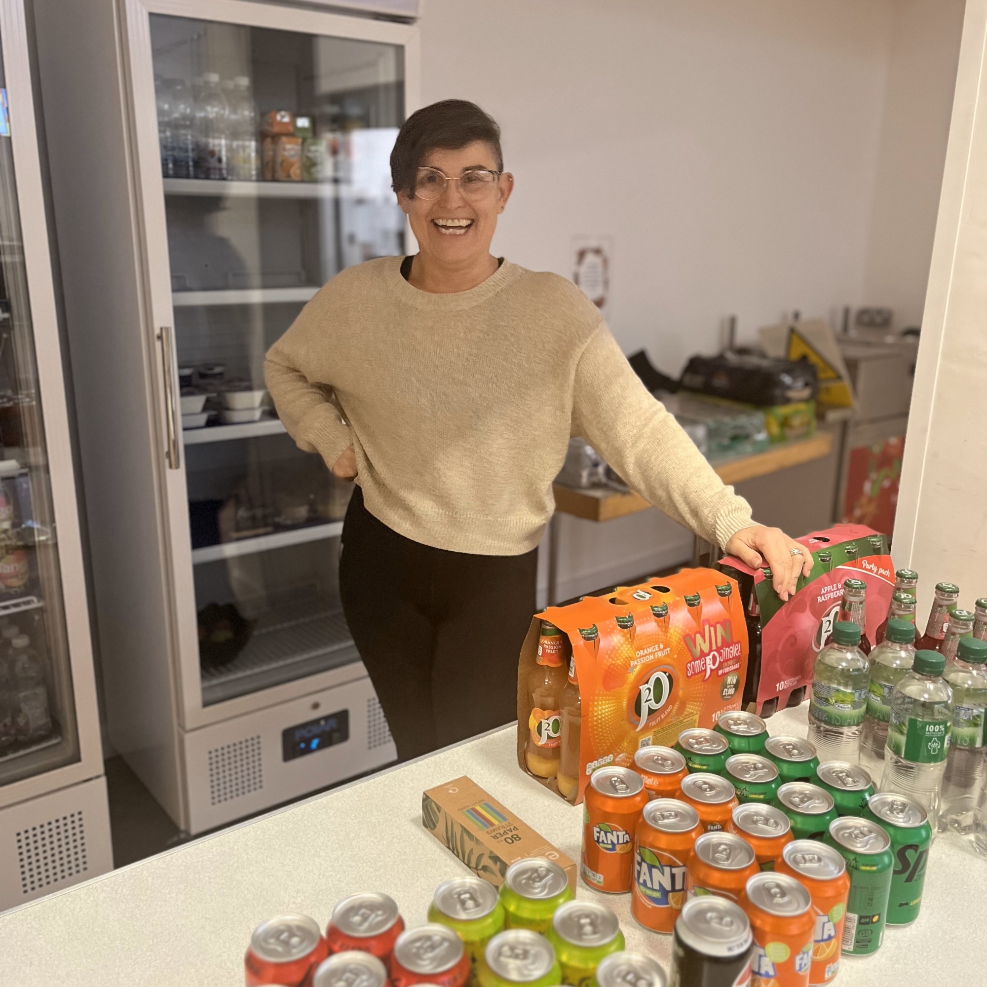 image of a women with short hair and a big smile, she is standing behind a table full of cans and bottles of drinks, the women is leaning on a box of the bottles of drink.  Behind  the women there  are fridges filled with drinks and boxes and a table