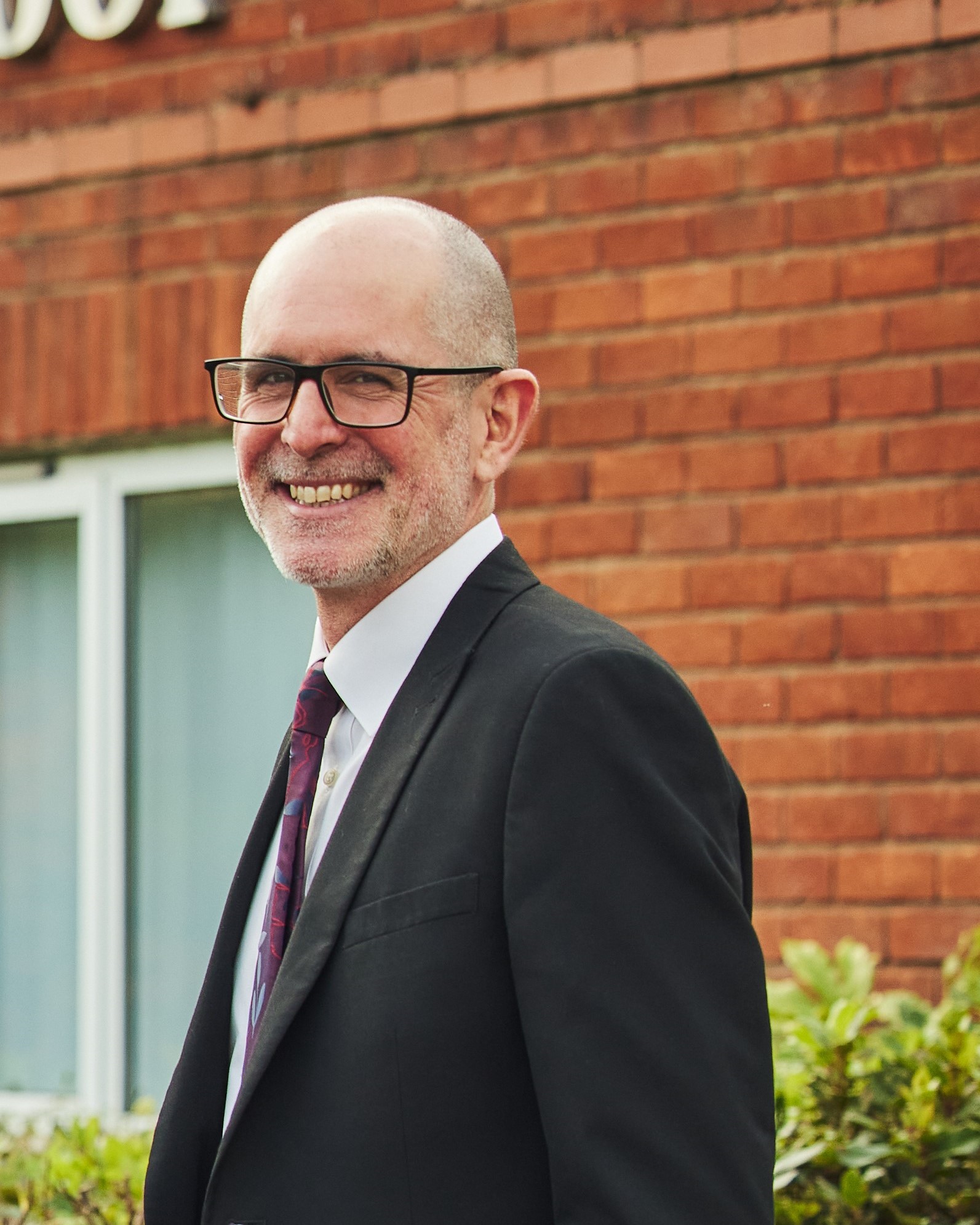 Image of a man standing to the side, looking at the camera smiling with a building in the background