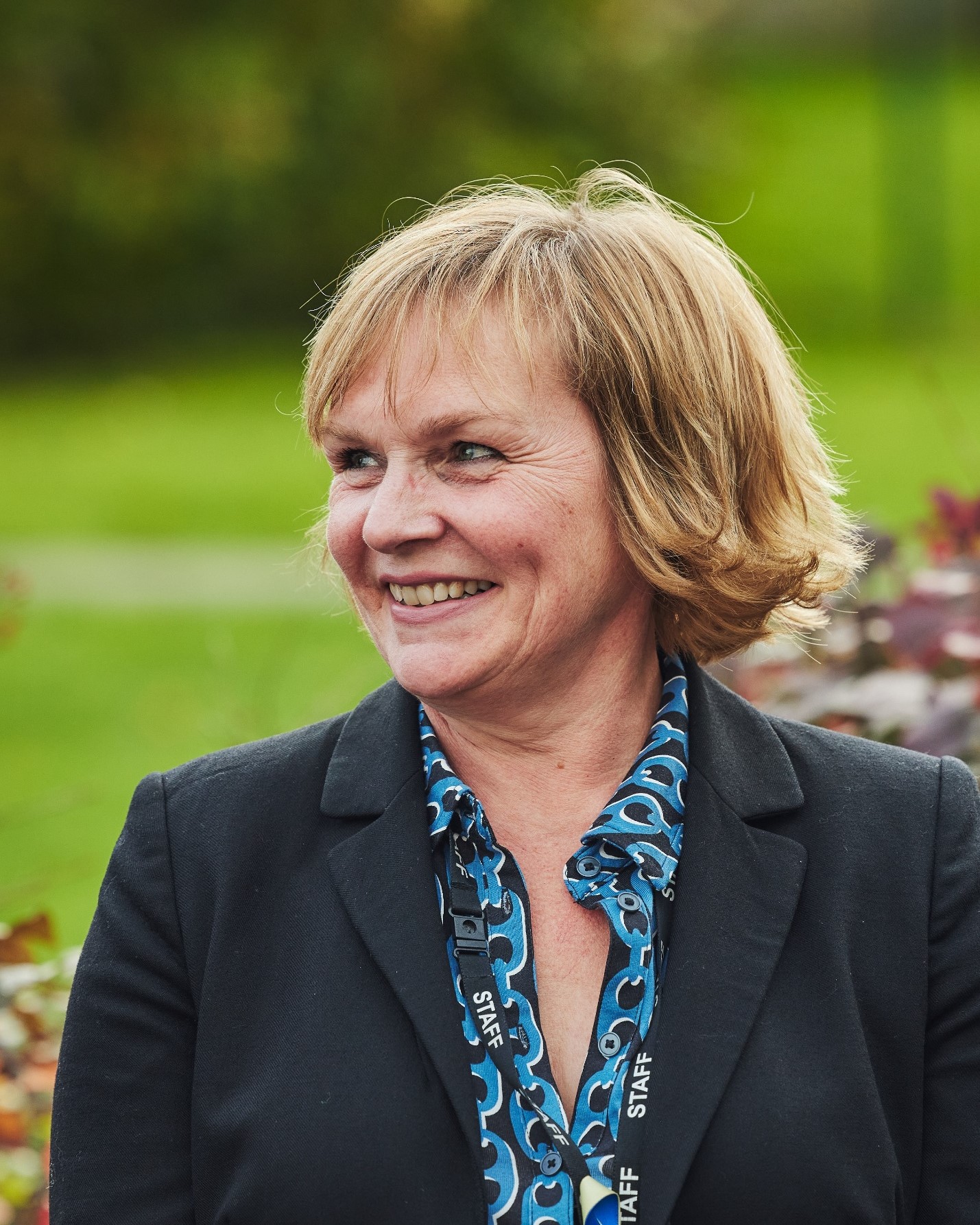 Image of a women smiling looking away from the camera, with a green field and some bushes behind her