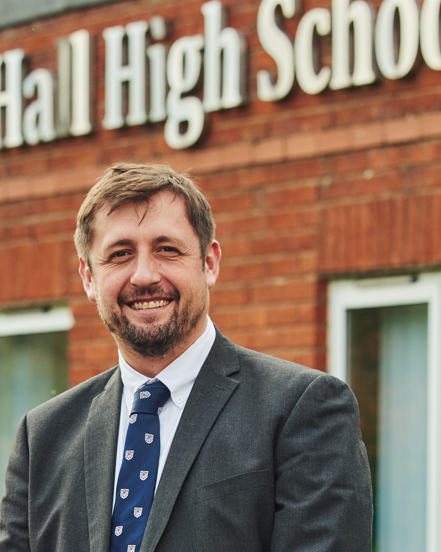 Picture of a man smiling at the camera, with a building and part of the the school name in the background