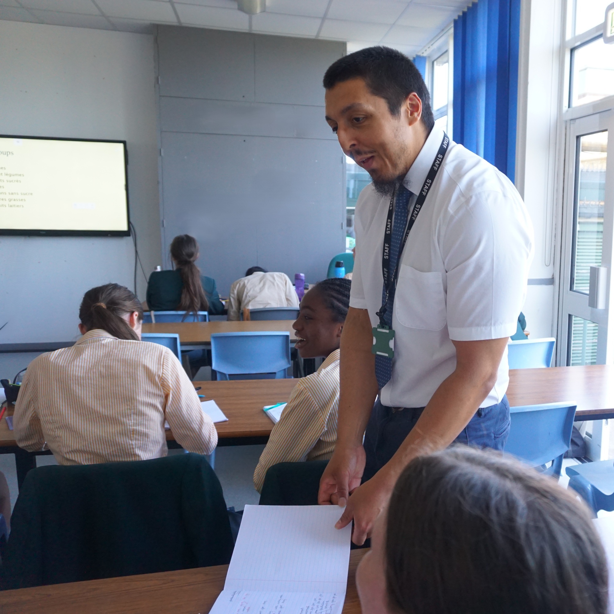 Picture of a male teacher standing talking to students  in a classroom. The female students are sitting in rows working and he is standing in between two rows